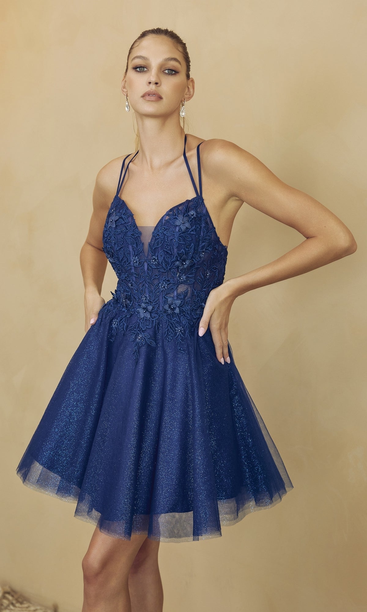 A woman models the Glitter-Tulle Short A-Line Homecoming Dress 864 in navy blue, featuring floral embroidery and a glitter-tulle skirt, set against a beige background.